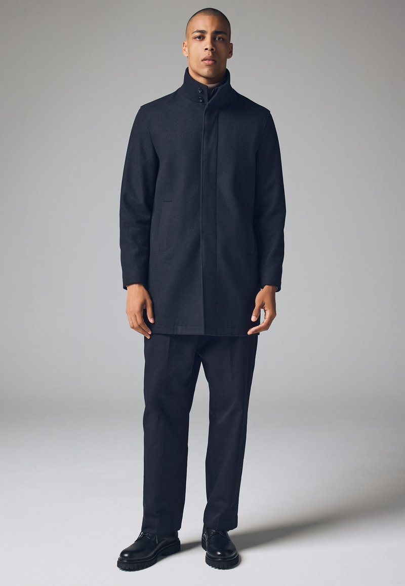 Zwarte wollen jas met een hoge kraag, lange mouwen en twee voorzakken, gecombineerd met een donkere broek en zwarte veterschoenen.