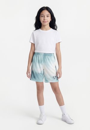 Jeune fille aux longs cheveux foncés, portant un t-shirt blanc, un short bleu et blanc, des chaussettes blanches et des baskets blanches, se tenant devant un fond uni.