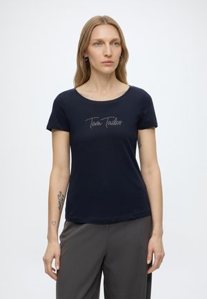 Kobieta o długich blond włosach, ubrana w granatowy t-shirt marki Tom Tailor i szare spodnie, stojąca na tle jednolitego jasnego tła.