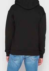 Czarna bluza z kapturem z przestronnym kapturem i ściągaczem na dole. Nosi się ją na jasnoniebieskie dżinsy z wzorem kieszeni.