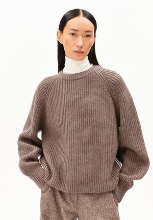 Pull en tricot côtelé marron avec un col rond et des épaules tombantes. Porté sur un col roulé blanc, avec une coupe décontractée et un motif texturé.