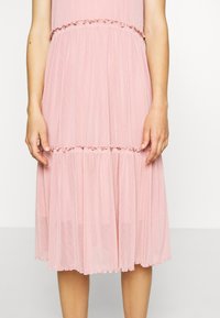 Różowa sukienka w odcieniu różanym z warstwowym krojem, ozdobiona falbankami na wysokości talii i u dołu. Wykonana z lekkiego, przezroczystego materiału o zwiewnej fakturze.