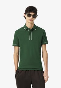 Polo shirt verde con piping bianco, realizzato in tessuto morbido. Presenta un colletto, maniche corte e un piccolo logo ricamato sul petto.