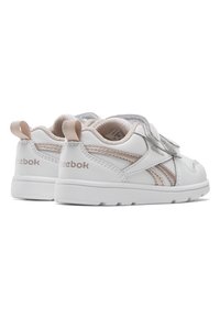 Vita Reebok-sneakers med beige detaljer, med texturerade paneler, kardborreband och en gummisula för bra grepp. Bakom finns en dragflik.