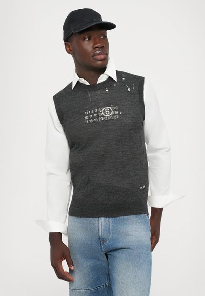 Uomo con un berretto nero, camicia bianca, maglione senza maniche grigio scuro con numeri e jeans di denim, che guarda di lato su uno sfondo bianco.