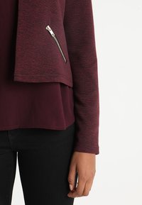 Veste bordeaux en tissu texturé, au design court, avec une poche avant zippée. Portée sur un haut sombre, associée à un pantalon noir.