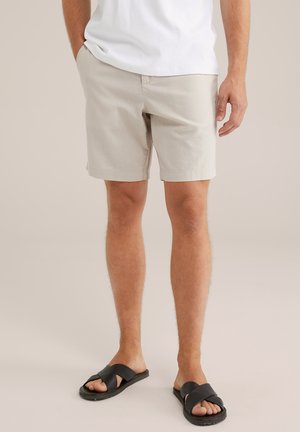 Beige cotton shorts with a straight cut, front pockets, and a flat waistband, paired with black slide sandals.