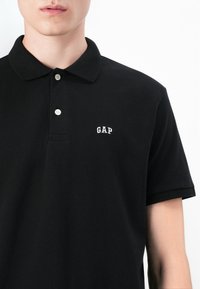 Polo shirt noir avec un col classique, deux boutons et un logo « GAP » blanc sur la poitrine. Fabriqué en tissu en coton texturé.