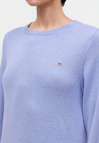 Femme portant un pull en maille violet clair avec un petit emblème blanc, rouge et noir sur la poitrine, montrée des épaules à la partie supérieure de la poitrine.