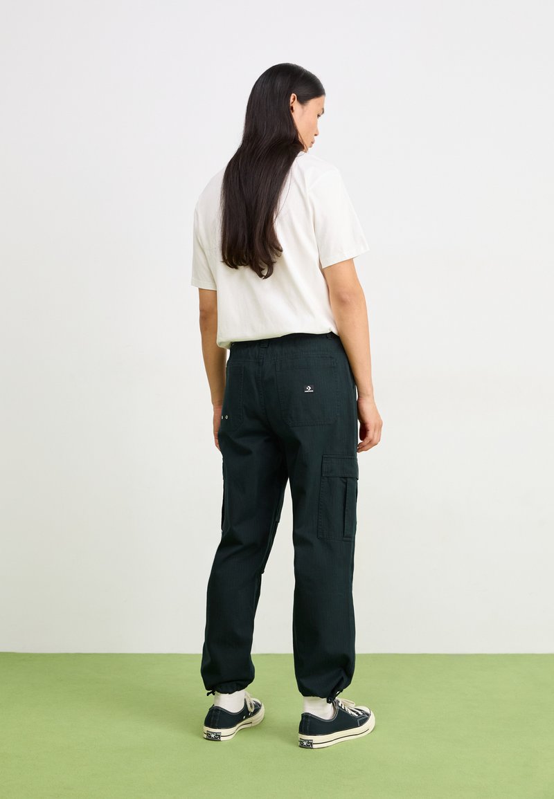 Pantalons cargo verts foncés avec poches latérales, chevilles resserrées et coupe décontractée, associés à un t-shirt blanc à manches courtes et des baskets noires.
