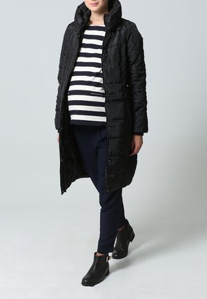 Sort, quiltet knælængdejakke med høj krave, båret over en stribet top og mørke bukser. Har lynlåslukning og en figursyet pasform.