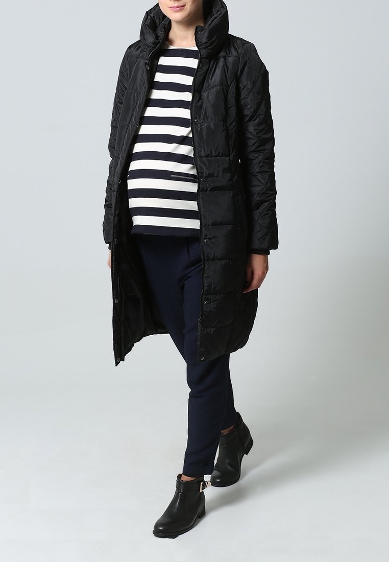 Zwarte, doorgestikte jas tot op de knie met een hoge kraag, gedragen over een gestreept topje en donkere broek. Voorzien van ritssluiting en een getailleerd ontwerp.