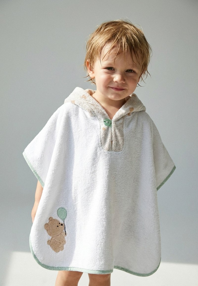 Jeune enfant aux cheveux mouillés portant un poncho à capuche blanc avec un motif d'ours en peluche tenant un ballon vert sur le côté.