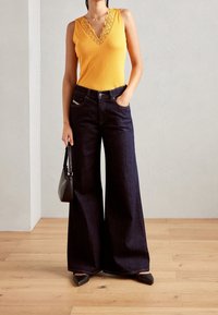 Gelbes ärmelloses Top mit Spitzenbesatz, kombiniert mit dunklen weiten Jeans. Die Accessoires umfassen eine kleine schwarze Handtasche und spitze Ballerinas.