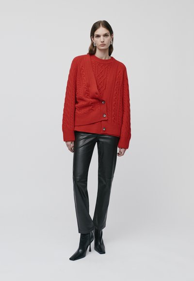 Raudonas megztinis su nertinėmis zigzagais, su mygtukais ir giliais V formos iškirpimu, derinamas su juodais odiniais kelnėmis ir aukštakulnėmis aulinėmis bateliais.