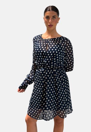 Donna che indossa un vestito blu navy con pois bianchi, maniche lunghe e vita arricciata, in piedi davanti a uno sfondo chiaro e uniforme.