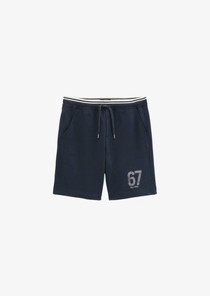 Giovane uomo in piedi che indossa una maglietta bianca, pantaloncini blu navy con il numero 67, sneakers bianche e una felpa blu navy appoggiata sulle spalle.
