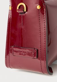Patent bordeaux handtas met gouden hardware accenten. Kenmerkt zich door een gestikt rand en een logo-label aan de zijkant. Gladde, glanzende textuur.
