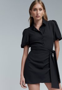 Vestido negro abotonado con mangas abullonadas, cintura con cinturón y diseño cruzado. Tela lisa con textura suave y dobladillo recto.