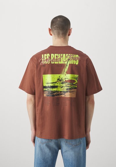 T-shirt en coton marron avec un design graphique coloré au dos, comprenant du texte et une illustration de voilier, associé à un jean en denim bleu clair.