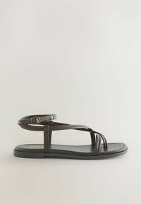 Tmavohnedá plochá sandála s tenkými remienkami, prackou na členku a minimalistickým otvoreným dizajnom prstov na svetlom pozadí.
