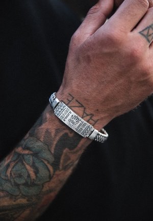 Zilveren armband met gegraveerde ontwerpen, gevlochten accenten en een rechthoekige centertool met een oogmotief. Draag je op een getatoeëerde pols.