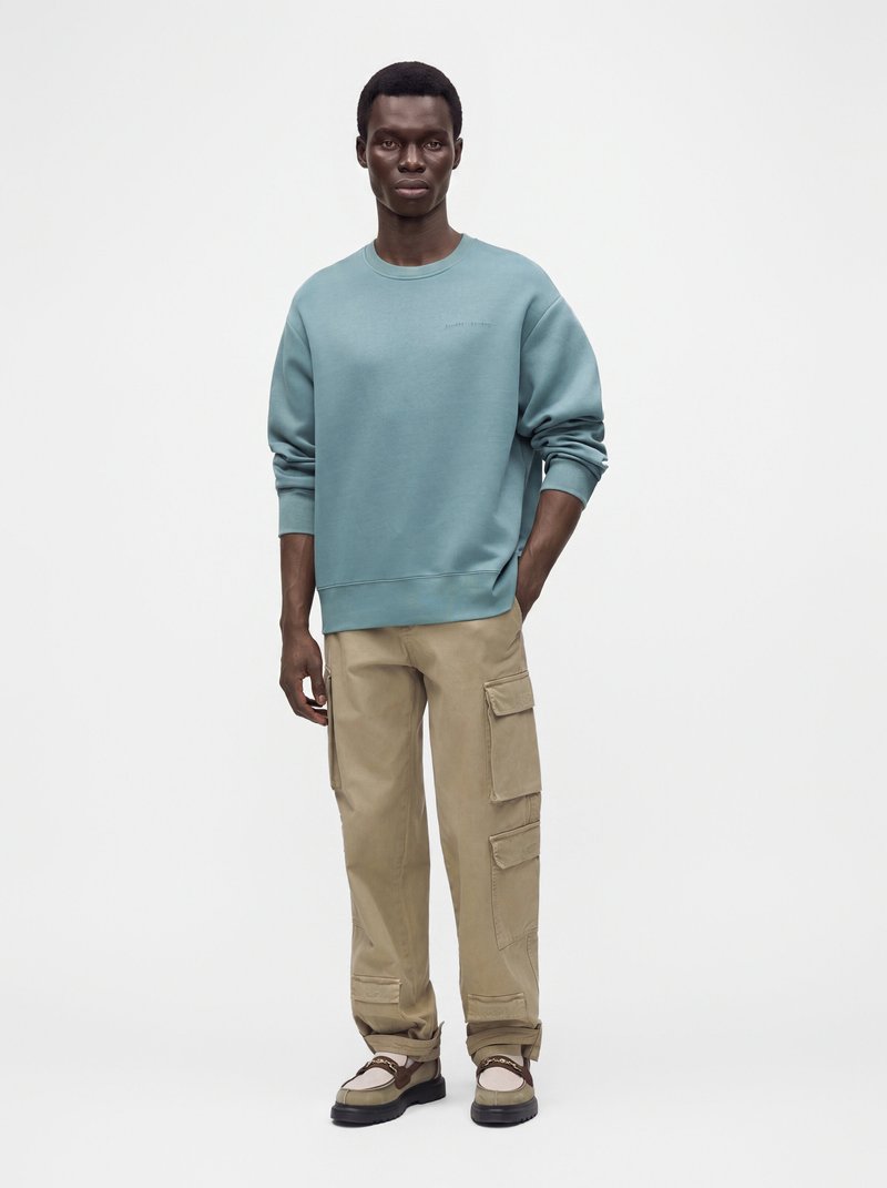 Homme debout portant un sweat-shirt bleu à col rond, un pantalon cargo beige et des chaussures beige et marron sur un fond blanc uni.