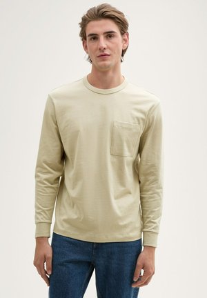 Langärmliges, hellbeiges T-Shirt aus weichem Stoff, mit rundem Halsausschnitt und einer linken Brusttasche, kombiniert mit blauen Jeans.