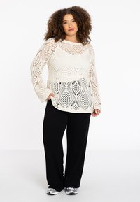 Gehaakte trui in crème met een ruitpatroon, gecombineerd met zwarte wijde broek en grijze sneakers. Krullend haar dat het gezicht omlijst.