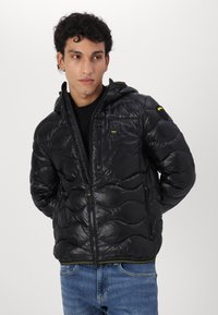 Giubbotto imbottito nero con finitura lucida, modello trapuntato a onde, chiusura frontale con zip e cappuccio. Include una piccola tasca con zip sul petto.