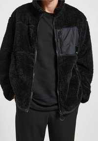 Fleece'owa kurtka w kolorze czarnym z teksturowanym wykończeniem. Posiada zasuwaną nylonową kieszeń na piersi. Zamek z przodu oraz luźny krój. Długie rękawy.