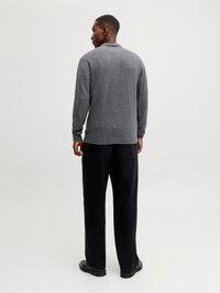 Mann, von hinten, trägt einen grauen Pullover, schwarze Hosen mit weitem Bein und schwarze Schuhe, steht vor einem schlichten hellen Hintergrund.