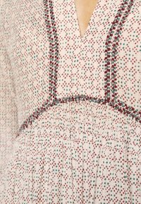 Tissu crème pour robe avec petits motifs en losange rouges et verts et coutures décoratives rouges et vertes le long des coutures et de l'encolure.