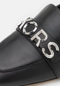 Scarpa slip-on nera in pelle con una fascia in metallo argentato con logo e dettagli cuciti lungo i bordi per un'aggiunta di texture e design.