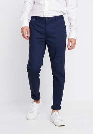 Homme portant un chino bleu marine slim, une chemise blanche à pois et des baskets blanches, debout sur un fond blanc.