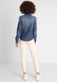 Dunkelblaues Denimhemd mit spitzem Kragen, langen Ärmeln mit Knopfmanschetten und abgerundhem Saum. Kombiniert mit hellen, ausgefransten Jeans.