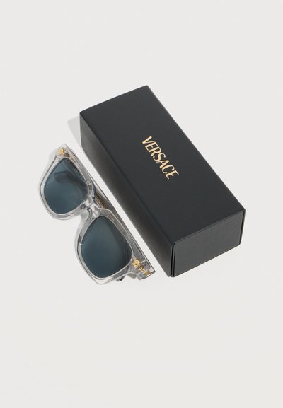 Γυαλιά ηλίου Versace με καθαρό πλαίσιο και σκούρους φακούς, τοποθετημένα δίπλα σε μαύρη θήκη με το λογότυπο Versace πάνω σε λευκή επιφάνεια.