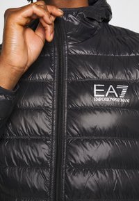 Svart pufferjacka med huva, med dragkedjestängning och horisontell sömnad. Logotyp "EA7 EMPORIO ARMANI" på bröstet. Slät textur.