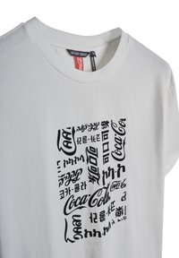 Witte T-shirt met een relaxte pasvorm, voorzien van zwarte Coca-Cola-logo's in verschillende talen en Antony Morato branding op de kraag en onderrand.