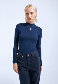 Marineblauses mit langen Ärmeln und hohem Kragen, aus glattem Stoff, kombiniert mit dunklen, hoch getäuschten Hosen mit Knopf- und Taschendetails.