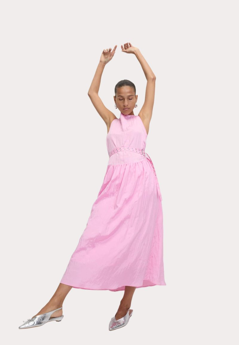 Robe maxi rose avec un col halter, tissu texturé et taille cintrée. Chaussures argentées métalliques à bout pointu et design minimaliste.