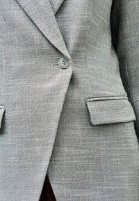 Blazer gris texturé avec une fermeture à un seul bouton et des poches à rabat, présenté en vue rapprochée.
