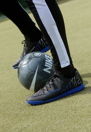 Person iført sort-hvide bukser kontrollerer en sort og blå Nike fodbold på en grøn kunstgræsbane.