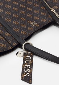 Sac à main Guess à motifs noirs et bruns avec fermeture éclair argentée, bandoulière noire et étiquette attachée portant "GUESS" en lettres argentées.