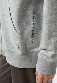 Szara bluza z miękką fakturą, z żebrowanymi ściągaczami na mankietach i dole, kieszeniami bocznymi oraz nadrukowaną metką z napisem "ZAPROJEKTOWANE DLA LUDZI".