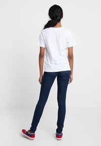 Wit katoenen T-shirt, gecombineerd met donkerblauwe skinny jeans. Schoenen hebben een rood-blauw ruitpatroon en stevige rode accenten.