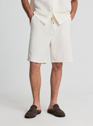 Uomo che indossa pantaloncini color avorio e camicia abbinata con le mani nelle tasche, abbinato a scarpe slip-on marrone scuro.