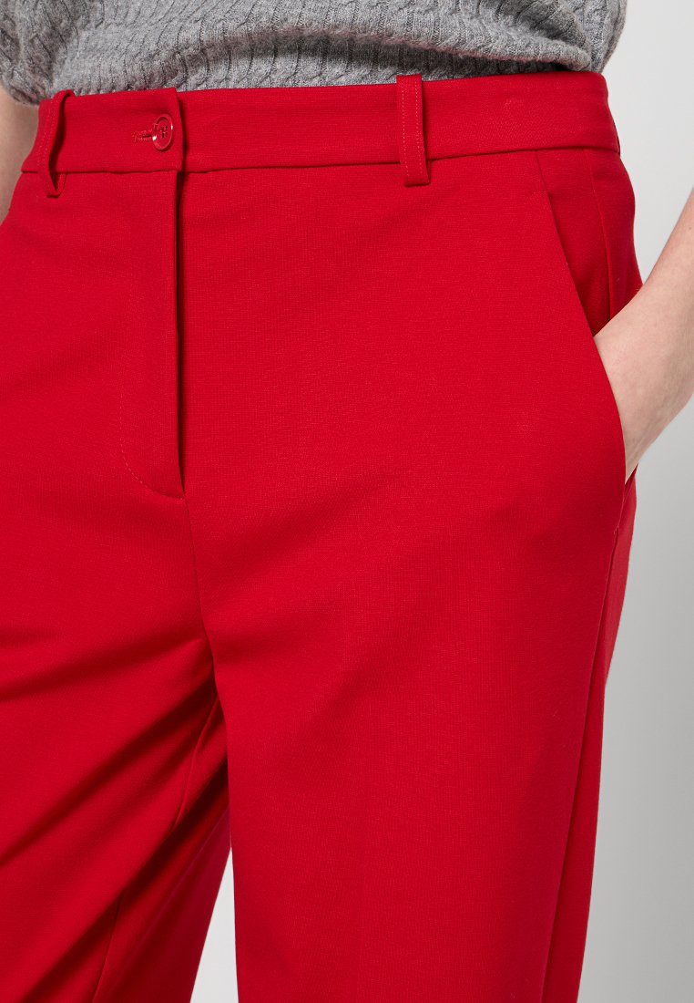 Pantaloni tailleur rossi con una texture liscia, caratterizzati da un fronte piatto, tasche laterali e passanti per cintura. Il tessuto ha una leggera lucentezza.