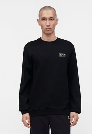 Mann trägt schwarzen Sweatshirt und Hose mit EA7 Emporio Armani-Logo auf der Brust und an der Hose vor einfachem weißen Hintergrund.