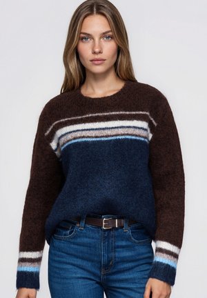 Une femme porte un pull à color-blocking, avec des manches marron, un corps bleu marine, des détails rayés et un jean bleu, sur un fond uni.